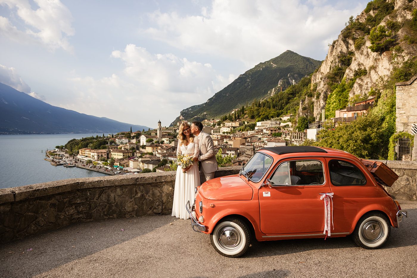 Coppia tedesca in posa con una Fiat 500 d&rsquo;epoca durante il loro matrimonio a Limone sul Garda, con il borgo e le montagne sullo sfondo.