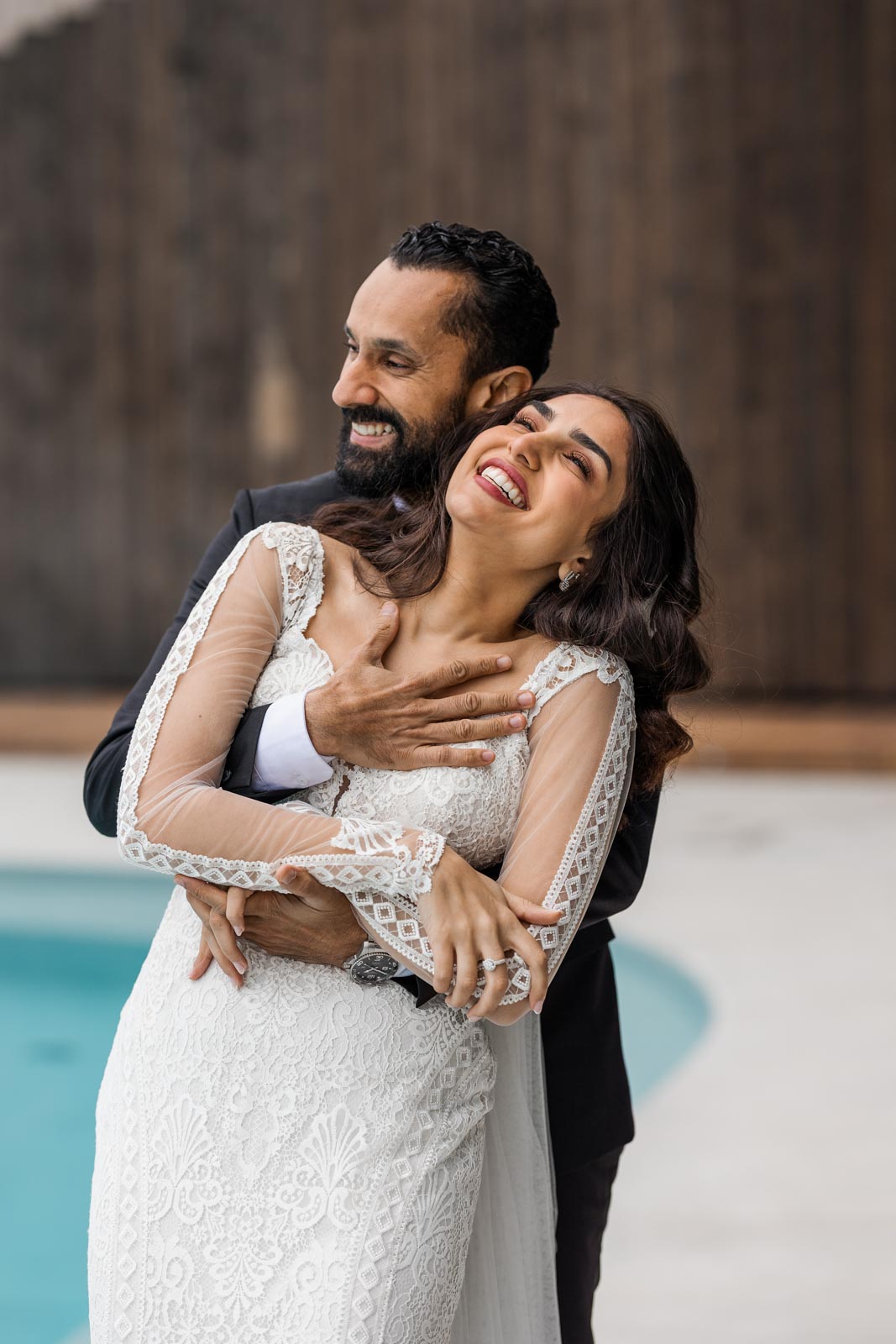 Bride laughing naturally in her groom’s arms during a relaxed wedding moment by the pool.