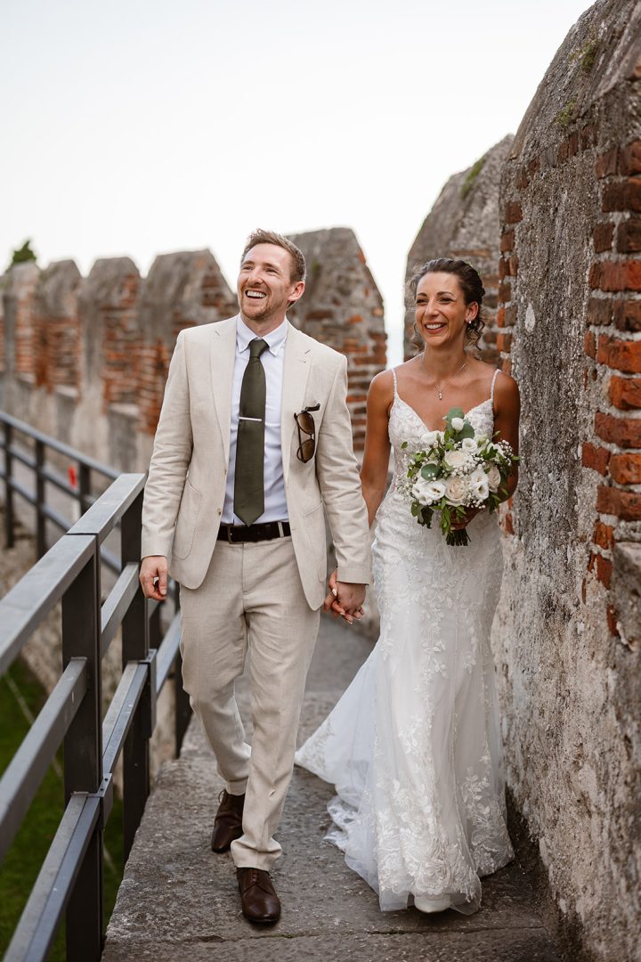Bride and groom walking hand in hand laughing on the walls of Castello di Malcesine, Lake Garda — destination wedding photography Italy